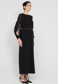 Czarna maxi sukienka z długimi, przezroczystymi rękawami i wycięciami. Posiada podkreśloną talię z cienkim paskiem, stylizowana z czarnymi szpilkami.