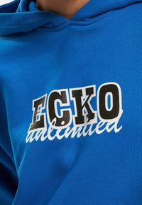Modrá bavlnená mikina s kapucňou, na prednej strane má grafiku s nápisom "ECKO" v tučných čiernych a bielych písmenách, s nápisom "unlimited" v kurzíve pod ním.