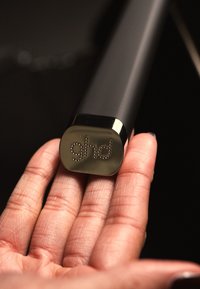 Handtag på svart hårverktyg som vilar i en hand, med en metallisk silverbas och präglad "ghd"-logotyp, slät textur och rundad form.