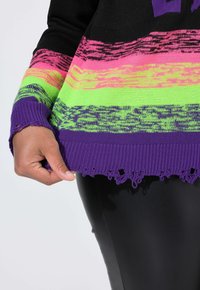 Sweatshirt en tricot noir avec des rayures horizontales colorées en rose, jaune et vert. Poignets côtelés violets et un ourlet à franges.