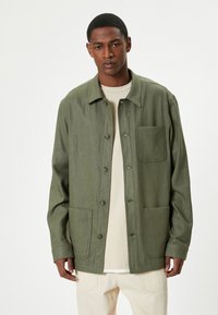 Mand iført en grøn knap-opsjacke med tre forlommer over en beige skjorte og lyse bukser imod en hvid baggrund.