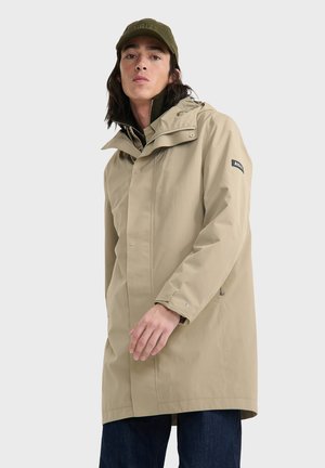 Giacca impermeabile beige con cappuccio, chiusura a bottoni e zip, due tasche laterali e patch con logo sulla manica. Design informale e luminoso.