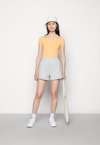 Πορτοκαλί κοντομάνικο μπλουζάκι με λευκό γραφικό, συνδυασμένο με γκρι σορτς με ελαστική μέση. Το μοντέλο φοράει λευκά αθλητικά παπούτσια και ένα λευκό καπέλο του μπέιζμπολ, κρατώντας μια τσάντα.