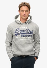 Felpa grigia in cotone con cappuccio regolabile, tasca a marsupio e testo grafico blu "SuperDry" sul davanti. Indossata con pantaloni scuri.