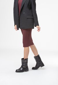 Stivaletti neri in pelle con punta arrotondata, fibbie laterali e suola in gomma testurizzata. Indossati con un abito fitted bordeaux e un blazer.