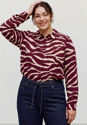 Femme souriante posant dans un chemisier à motifs bordeaux et crème et un jean foncé, une main reposant sur sa tête.