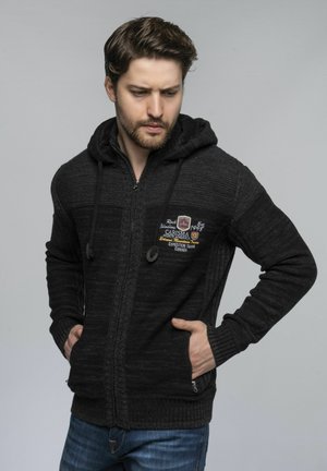Schwarze Zip-Hoodie mit Strickstruktur, Kordelzügen, zwei seitlichen Taschen und einem gestickten Logo auf der linken Brustseite.