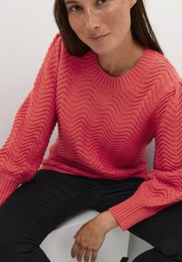 Coral knit sweater with wavy texture and ribbed cuffs, paired with black pants. Features a round neckline and loose fit.