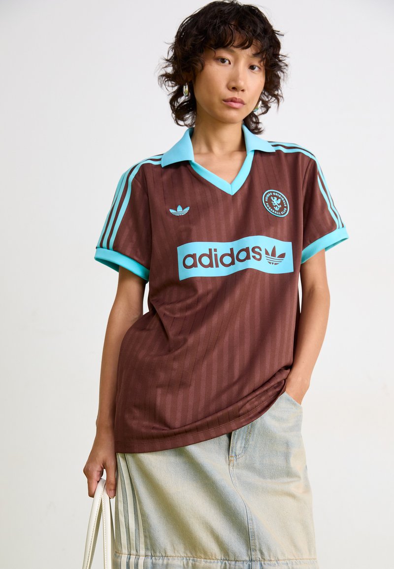 Camiseta deportiva marrón con rayas verticales, cuello y acentos turquesa. Presenta un gran logo de "adidas" en el frente y un emblema en el pecho.