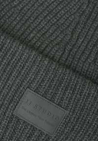 Donkergrijs ribgebreid materiaal met een zwart rechthoekig label dat is geperst met de tekst "JJ Studio Our Legacy Your Future."