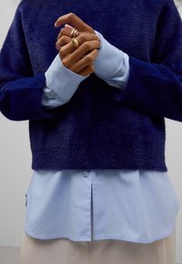 Marineblå, blød sweater lagdelt over en lyseblå skjorte med krave. De synlige hænder bærer tre guldringe. Glat stoftekstur.