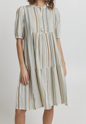 Kvinde iført hvid kjole i mellemlængde med lodrette beige, blå og brune striber, korte pufærmer og knapdetalje foran.