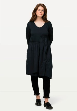 Abito nero con scollatura a V, maniche lunghe e dettagli in vita arricciati; abbinato a pantaloni neri con bordi in pizzo e scarpe nere.