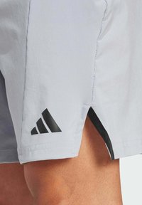 Los pantalones cortos Adidas de color gris claro presentan un logo negro en el lado izquierdo, con aberturas laterales en el dobladillo para mayor libertad de movimiento. Textura de tela suave.