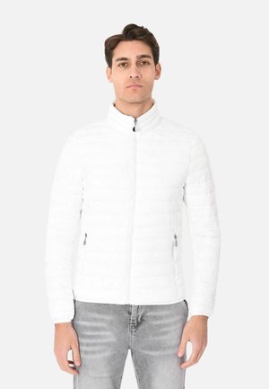 Giovane uomo che indossa una giacca trapuntata bianca con cerniera e jeans grigio chiaro, in piedi contro uno sfondo chiaro uniforme.