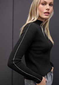 Czarny sweter z prążkowanej dzianiny z wysokim kołnierzem, z kontrastującymi białymi paskami po bokach. Model nosi go z jasnoszarymi dżinsami.