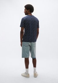 Μπλε ναυτικό T-shirt με λευκές πουά, λαιμόκοψη με ρεγκλάν; σετ με ανοιχτόχρωμα τζιν σορτς. Το μοντέλο στέκεται με την πλάτη στην κάμερα.