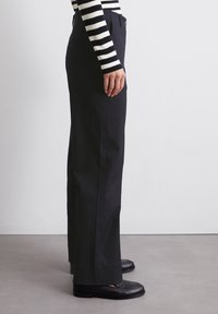 Pantalon noir taille haute avec une coupe droite. Confectionné en tissu texturé, il comprend des poches latérales et une fermeture à bouton. Associé à des mocassins noirs.