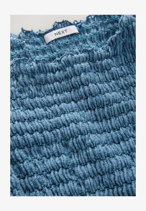 Textured blue fabric with a ruffled design and subtle sheen; includes a white label with the brand name "NEXT" near the top edge.