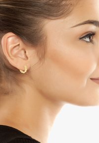 Goldene geometrische Creolen mit facettiertem Design, die am Ohrläppchen eines Models befestigt sind. Die Ohrringe haben eine polierte Oberfläche und kantige Formen.