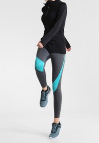 Černý dlouhý rukáv sportovní top s límcem; šedé legíny se tyrkysovými bočními panely; modré sportovní boty s texturovaným vzorem.