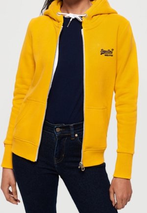 Osoba ubrana w jaskrawożółtą bluzę z kapturem na suwak, na koszulce granatowego koloru i ciemnoniebieskich dżinsach, stojąca z rękami swobodnie opuszczonymi wzdłuż ciała.