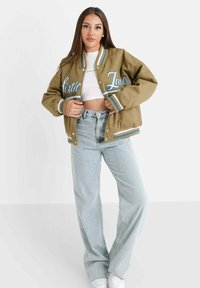 Bruine bomberjack met blauwe letters, geribbelde kraag en gestreepte accenten. Gekombineerd met lichtblauwe hoge taille jeans en witte sneakers.