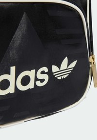 Czarna torba z tkaniny z białym logo „adidas” i motywem liścia koniczyny, posiadająca teksturowaną powierzchnię oraz złote okucia zamka.