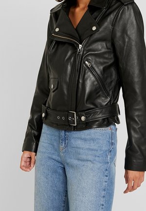 Sort læder bikerjakke med sølvfarvede lynlåse og bælte båret med lyseblå cowboybukser på en person mod en grå baggrund.