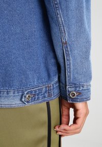 Veste en denim de couleur bleu clair, avec une coupe classique, des boutons pression en métal et des coutures contrastées sur les poignets.