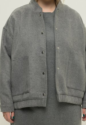 Personne portant une veste grise ample à boutons-pression sur une robe grise assortie, mains visibles avec un vernis à ongles vert foncé.