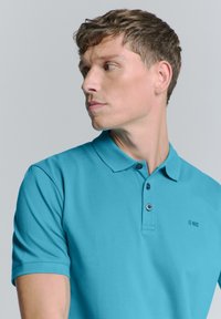 Turquoise poloshirt van zacht materiaal met een kraag-halslijn, drie knopen, korte mouwen en een klein zwart logo op de borst.