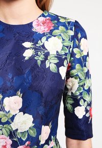 Tummansininen kukallinen mekko lyhyillä hihoilla, jossa on suuria vaaleanpunaisia ja valkoisia ruusuja sekä vihreitä lehtiä teksturoituneella kankaalla.