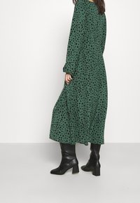 Grünes langärmliges Kleid mit schwarzen Pünktchen, fließender Form und seitlichen Taschen, kombiniert mit schwarzen kniehohen Blockabsatzstiefeln.