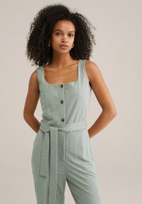 Ljusgrön jumpsuit med fyrkantig halsringning, framknappar och en knuten midjebälte. Har ett texturerat tyg och vida axelband.