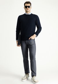 Navy gestrickter Pullover mit strukturiertem Muster, graue maßgeschneiderte Hose und beigefarbene Sneakers. Das Model hält eine schwarze Laptoptasche und trägt eine Sonnenbrille.