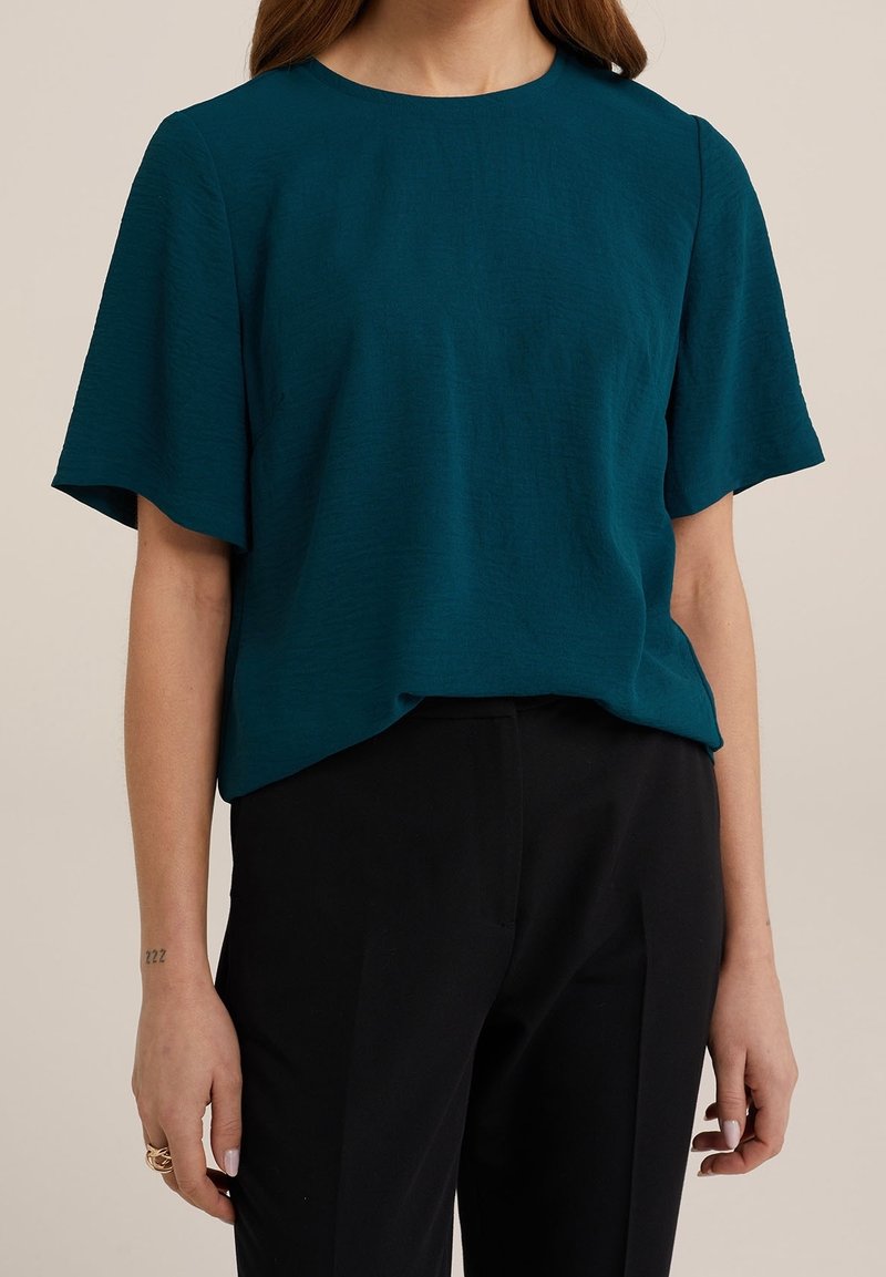 Teal kortärmad topp gjord av texturerad tyg, med rund halsringning och en avslappnad, minimalistisk design, kombinerad med svarta byxor.