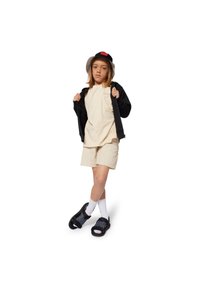 Child in beige t-shirt with pocket, cream shorts, black zip-up jacket, black bucket hat, white socks, and patterned black sandals.