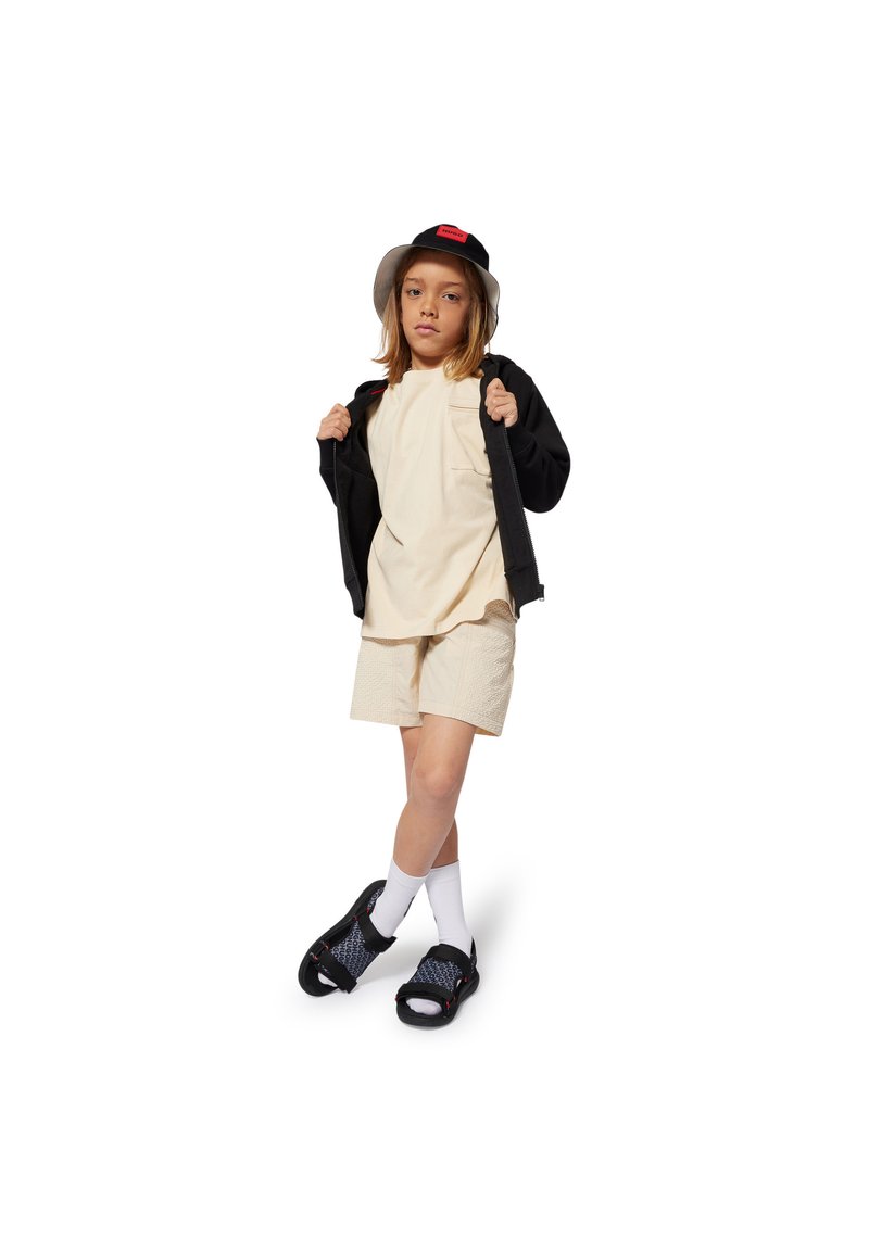 Child in beige t-shirt with pocket, cream shorts, black zip-up jacket, black bucket hat, white socks, and patterned black sandals.