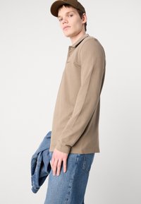 Camisa polo de mangas longas castanha com colarinho, apresentando um logótipo discreto. O modelo usa jeans de ganga azuis, segurando uma jaqueta azul.