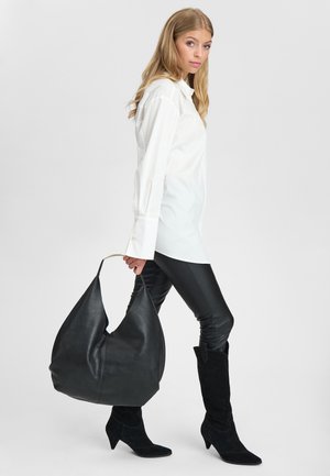 Kvinde går mod højre iført hvid skjorte med lange ærmer, sorte læderbukser og støvler, holder stor sort lædertaske med gyldent håndtag.