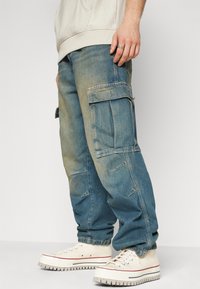 Ljusblå denim cargobyxor med flera fickor, avslappnad passform och elastiska muddar, kombinerat med off-white sneakers.