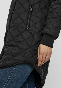 Veste noire matelassée avec une poche zippée, des poignets côtelés et un ourlet incurvé. Le tissu a une texture lisse avec des motifs en losange.