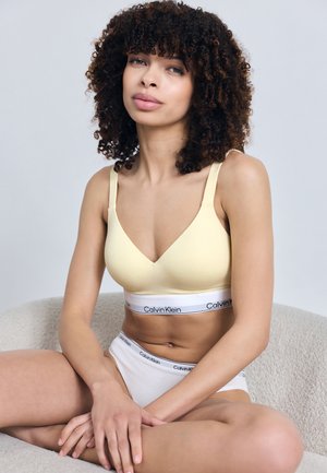 Ung kvinne med krøllete hår som sitter med korslagte ben iført en gul bralette og hvite Calvin Klein-undertøy mot en ensfarget bakgrunn.