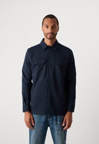 Man wearing a navy button-up shirt with two chest pockets and light blue jeans, standing against a plain light background.