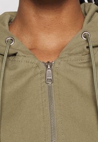 Olive green zip-up hoodie with a textured finish, featuring a silver zipper and metal eyelets for drawstrings.