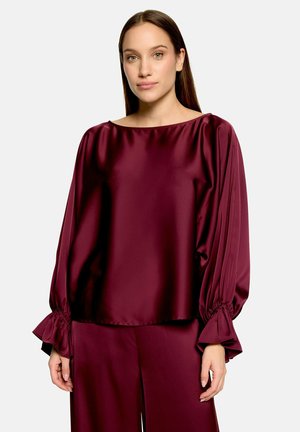 Femme portant un chemisier en satin bordeaux à manches longues avec poignets froncés et pantalon ample assorti sur un fond clair uni.