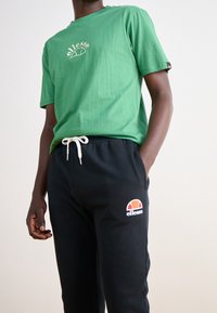 Grön bomullströja med en rundad logotypdesign på bröstet, kombinerad med svarta sweatpants som har en färgglad logotyp på låret.