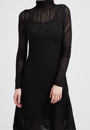 Robe noire en tricot avec manches longues, col montant, motifs texturés et jupe évasée. Présente des motifs de point complexes et une légère transparence.