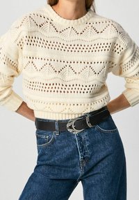 Krämfärgad stickad tröja med öppet mönster, kombinerad med högmidjade blå jeans och ett svart bälte med en silverfärgad utsmyckad spänne.